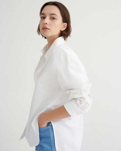 A person with short brown hair wears a Kowtow Season Shirt in white, styled in a relaxed fit with blue pants, standing against a plain white background and facing slightly sideways.