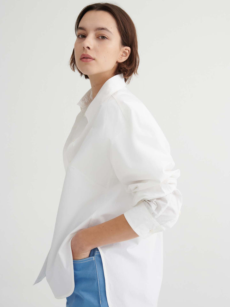 A person with short brown hair wears a Kowtow Season Shirt in white, styled in a relaxed fit with blue pants, standing against a plain white background and facing slightly sideways.