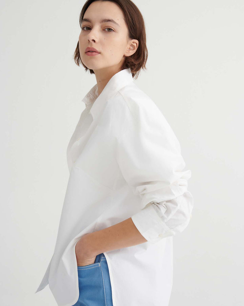 A person with short brown hair wears a Kowtow Season Shirt in white, styled in a relaxed fit with blue pants, standing against a plain white background and facing slightly sideways.