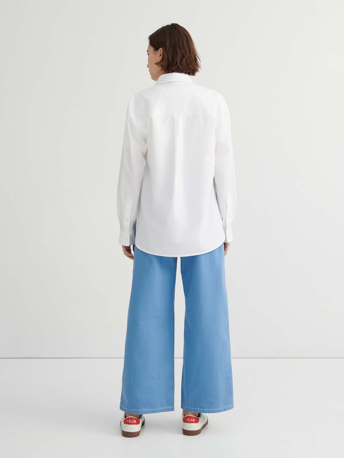 A person stands with their back to the camera, wearing the relaxed fit Season Shirt – White, paired with light blue wide-leg jeans and red and white sneakers against a plain white background.