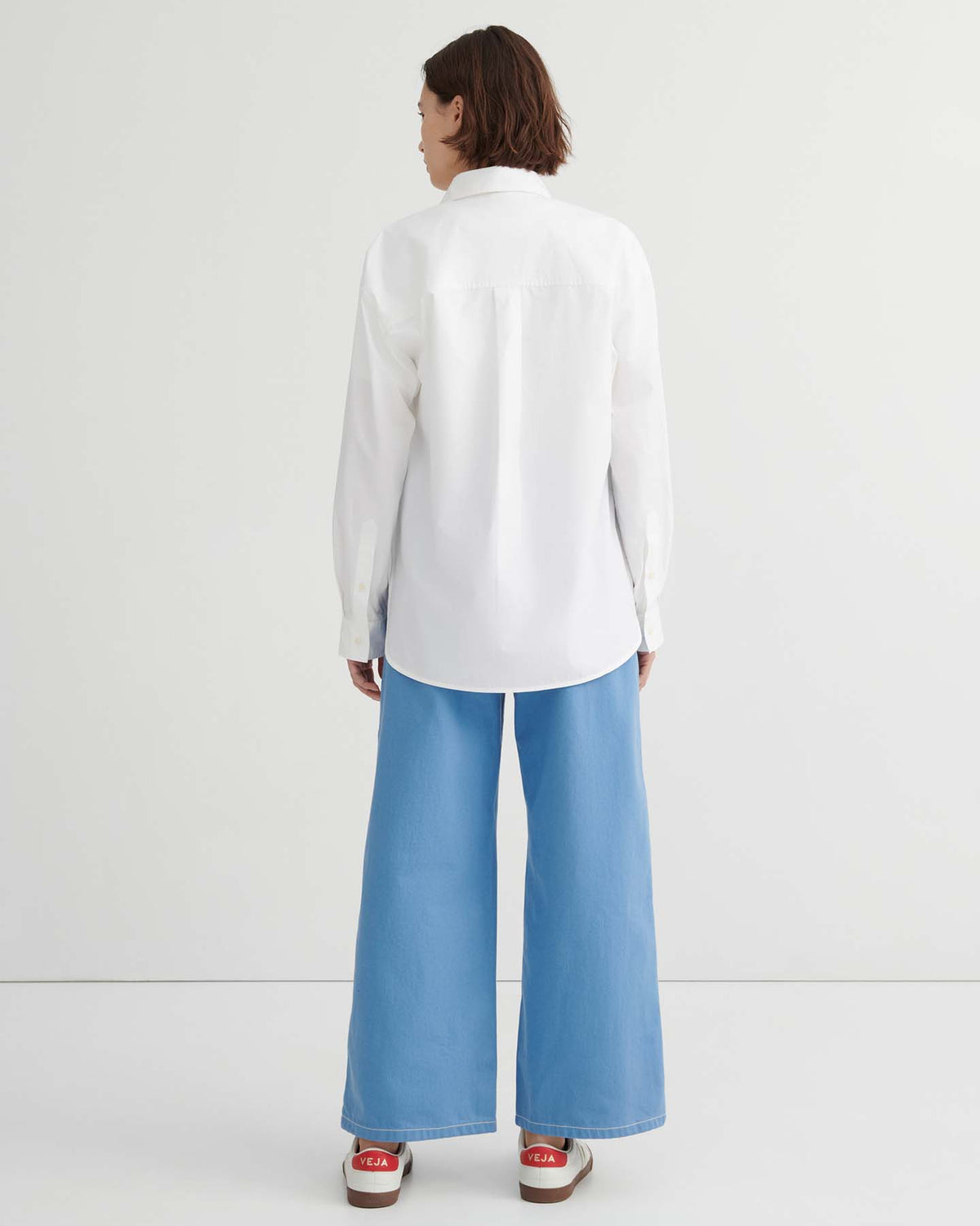 A person stands with their back to the camera, wearing the relaxed fit Season Shirt – White, paired with light blue wide-leg jeans and red and white sneakers against a plain white background.