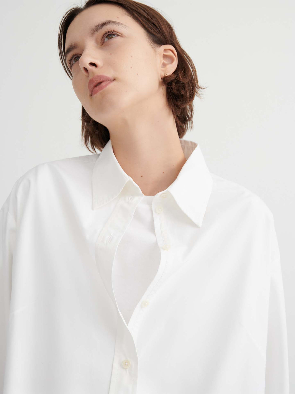 A person with short brown hair wears the Season Shirt – White by Kowtow in a relaxed fit, looking upward against a plain light background.