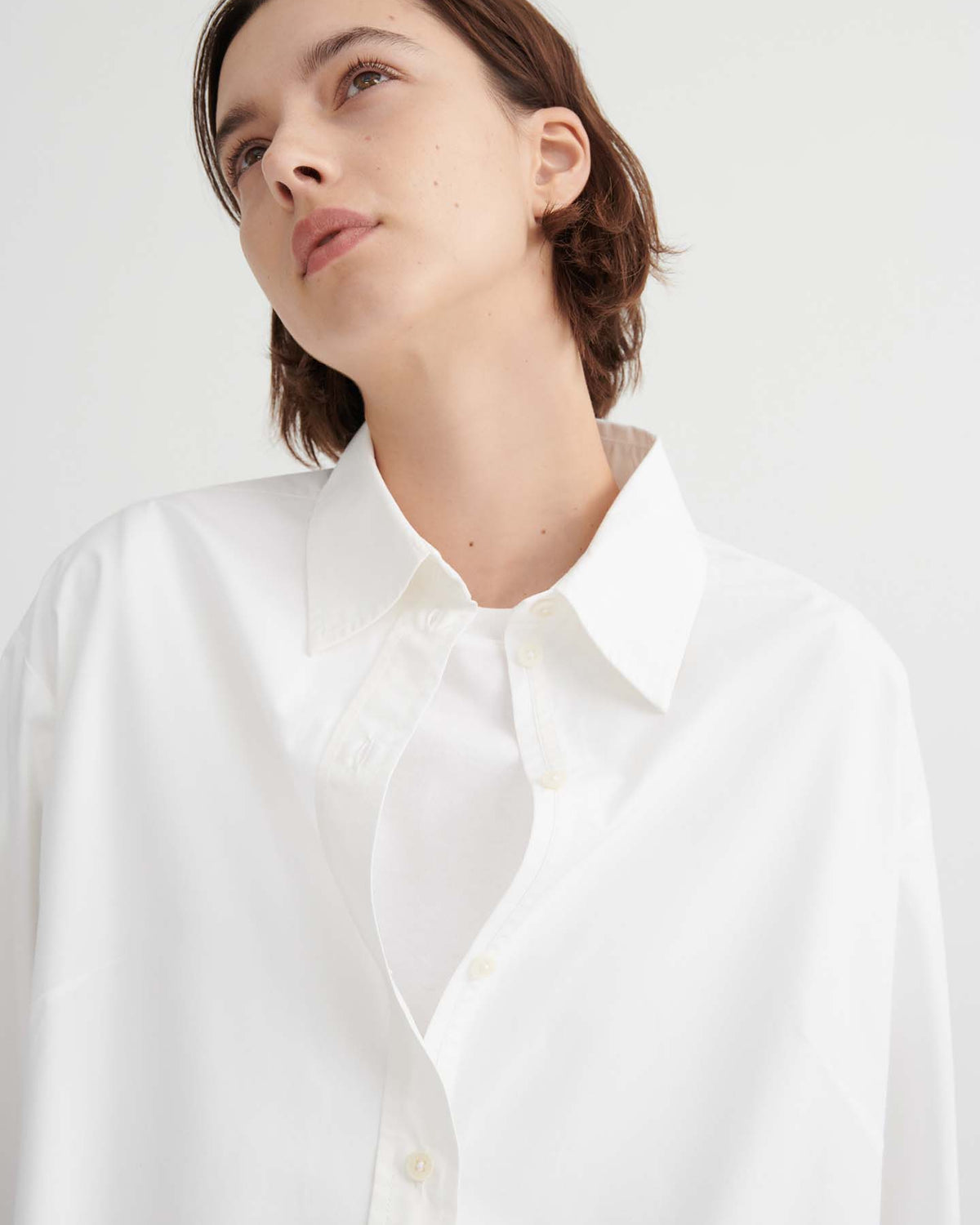 A person with short brown hair wears the Season Shirt – White by Kowtow in a relaxed fit, looking upward against a plain light background.