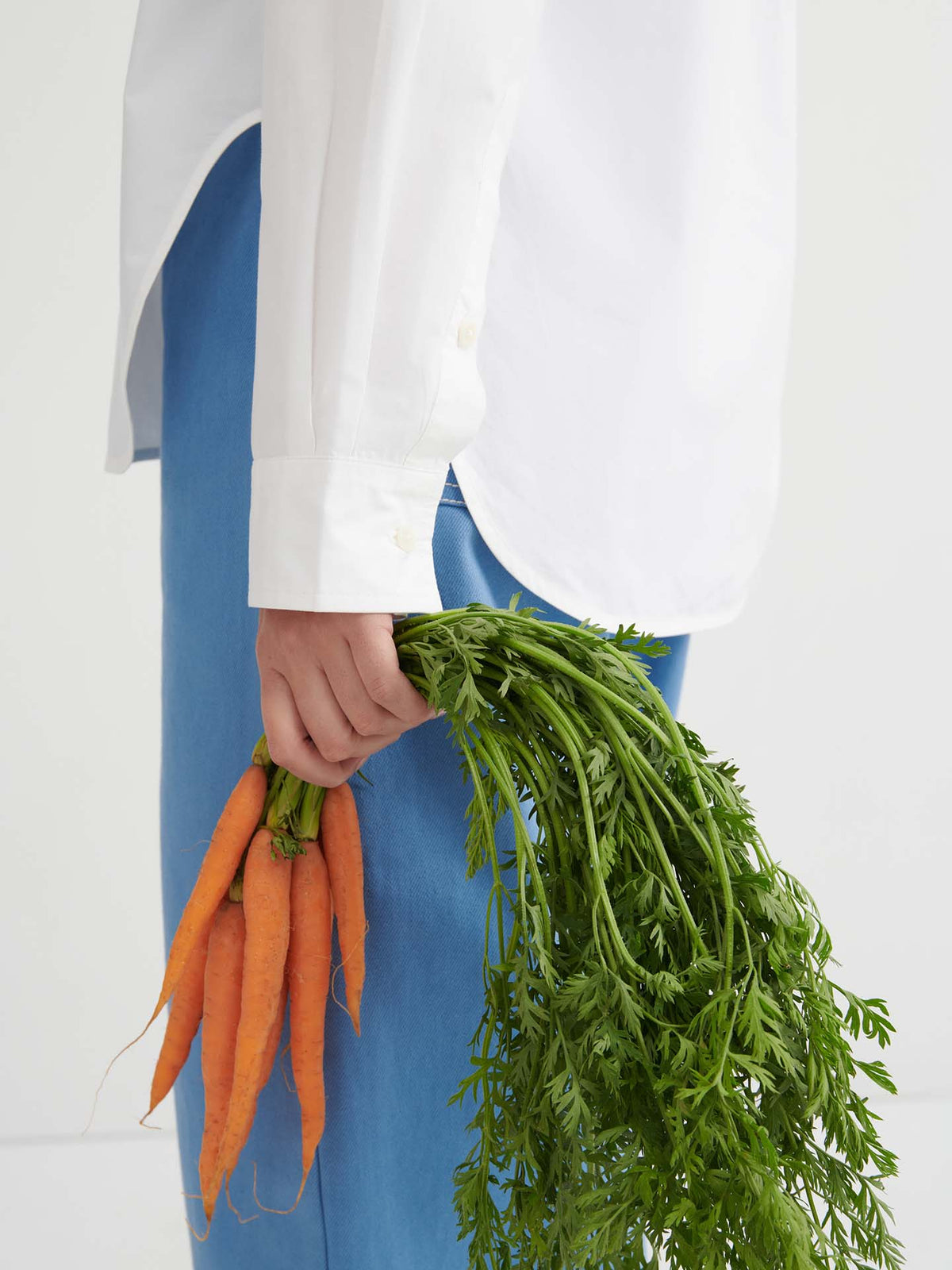 A person wearing the Season Shirt – White, styled in a relaxed fit and paired with blue pants, is holding a bunch of fresh carrots with green tops, shown from the side against a plain background.
