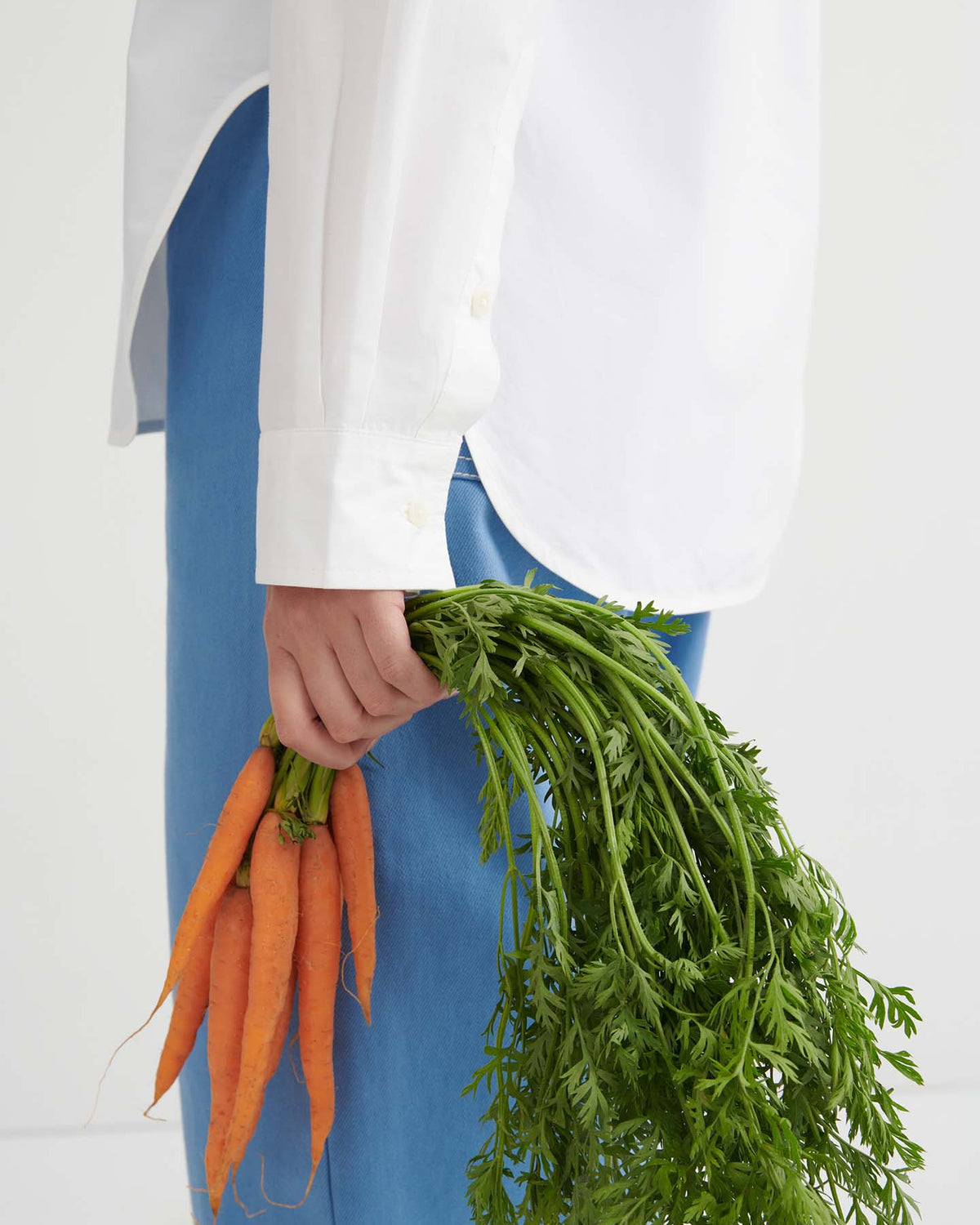 A person wearing the Season Shirt – White, styled in a relaxed fit and paired with blue pants, is holding a bunch of fresh carrots with green tops, shown from the side against a plain background.