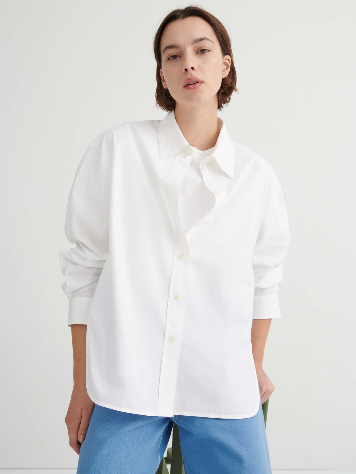 A person stands against a plain background wearing the loose-fitting White Season Shirt from Kowtow and blue pants, with one hand in their pocket, highlighting the shirt’s relaxed fit.