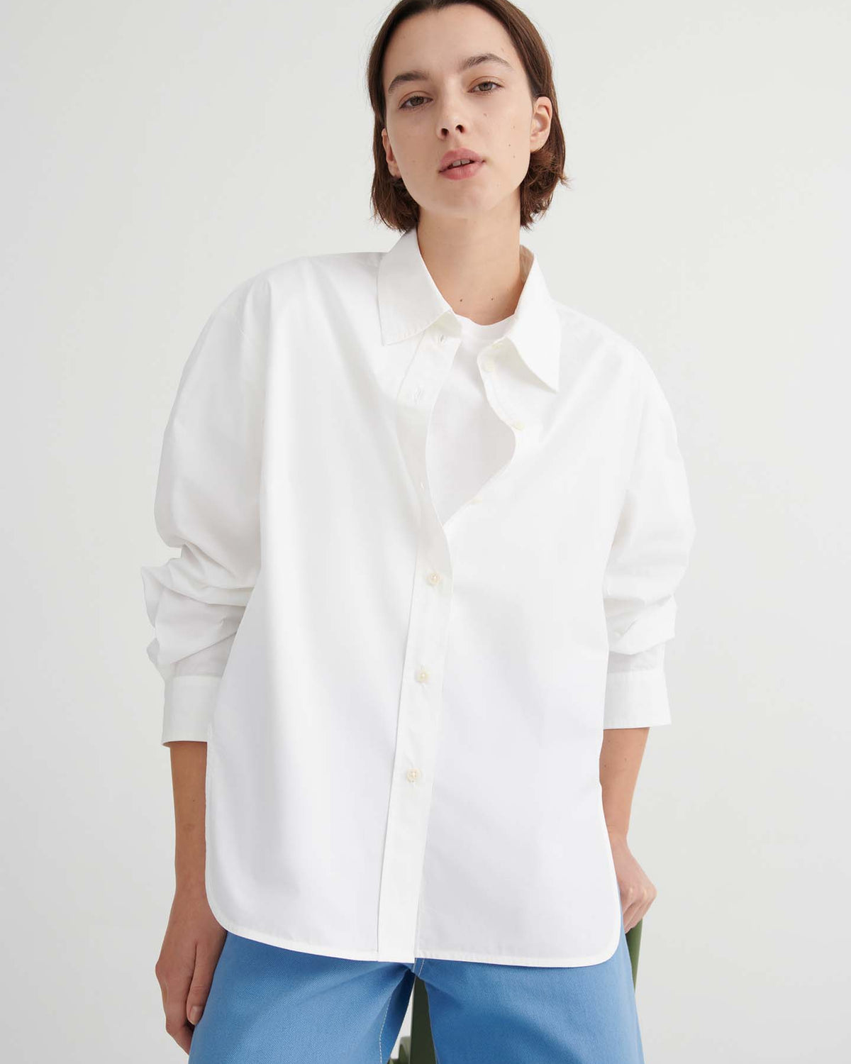 A person stands against a plain background wearing the loose-fitting White Season Shirt from Kowtow and blue pants, with one hand in their pocket, highlighting the shirt’s relaxed fit.