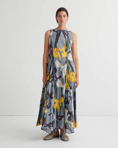 Person standing against a plain white background wearing the Signify Dress – Iris, a sleeveless, ankle-length piece featuring a bold yellow, white, and purple floral pattern. The garment’s standard fit offers ease of movement and pairs perfectly with flat sandals.