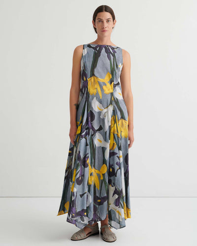 Person standing against a plain white background wearing the Signify Dress – Iris, a sleeveless, ankle-length piece featuring a bold yellow, white, and purple floral pattern. The garment’s standard fit offers ease of movement and pairs perfectly with flat sandals.