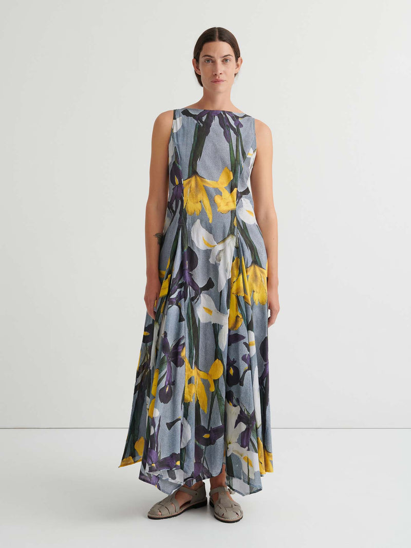 Person standing against a plain white background wearing the Signify Dress – Iris, a sleeveless, ankle-length piece featuring a bold yellow, white, and purple floral pattern. The garment’s standard fit offers ease of movement and pairs perfectly with flat sandals.