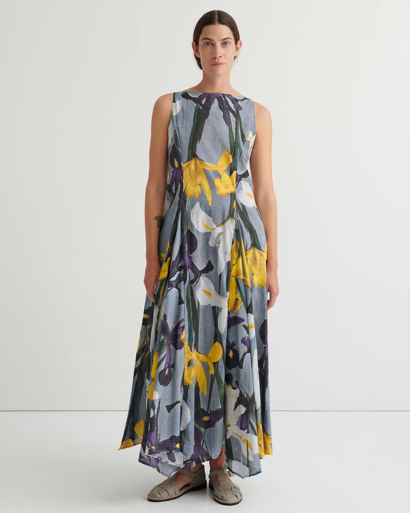 Person standing against a plain white background wearing the Signify Dress – Iris, a sleeveless, ankle-length piece featuring a bold yellow, white, and purple floral pattern. The garment’s standard fit offers ease of movement and pairs perfectly with flat sandals.