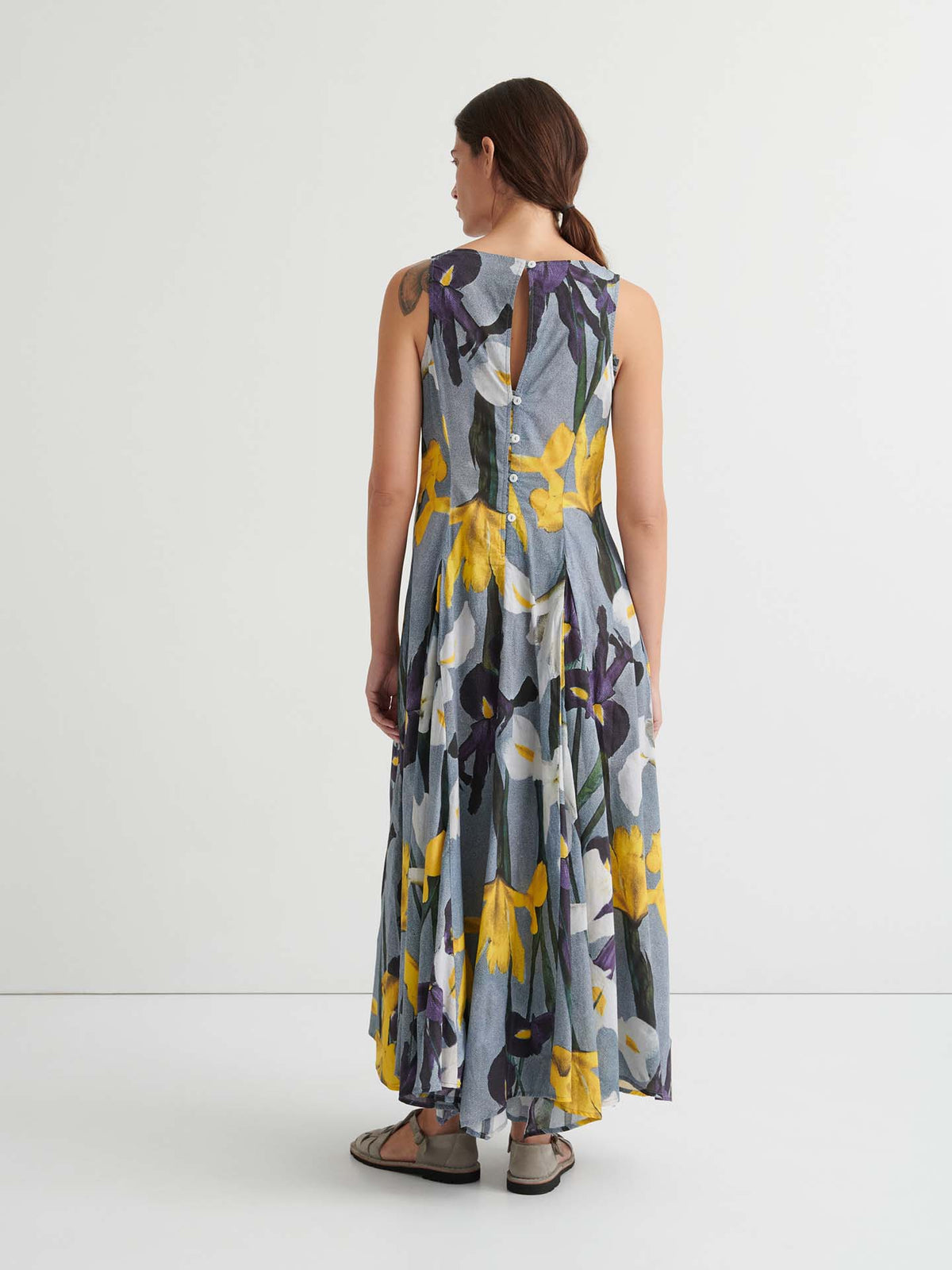 A person stands with their back to the camera, wearing the Signify Dress – Iris: a standard-fit, sleeveless, ankle-length dress adorned with a striking purple and yellow floral print and finished with a buttoned keyhole back. Carefully designed garment measurements offer a flattering silhouette from every angle.