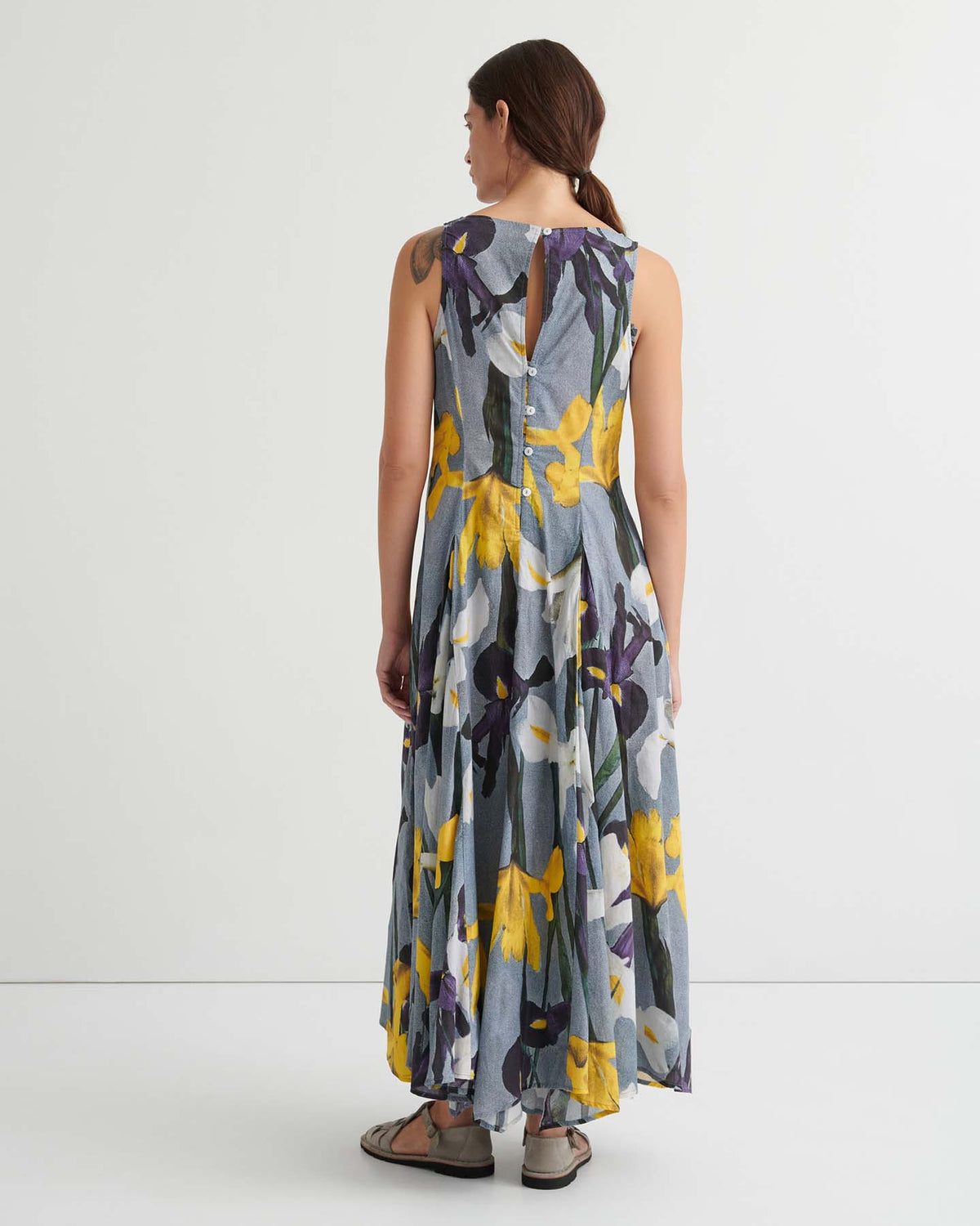 A person stands with their back to the camera, wearing the Signify Dress – Iris: a standard-fit, sleeveless, ankle-length dress adorned with a striking purple and yellow floral print and finished with a buttoned keyhole back. Carefully designed garment measurements offer a flattering silhouette from every angle.