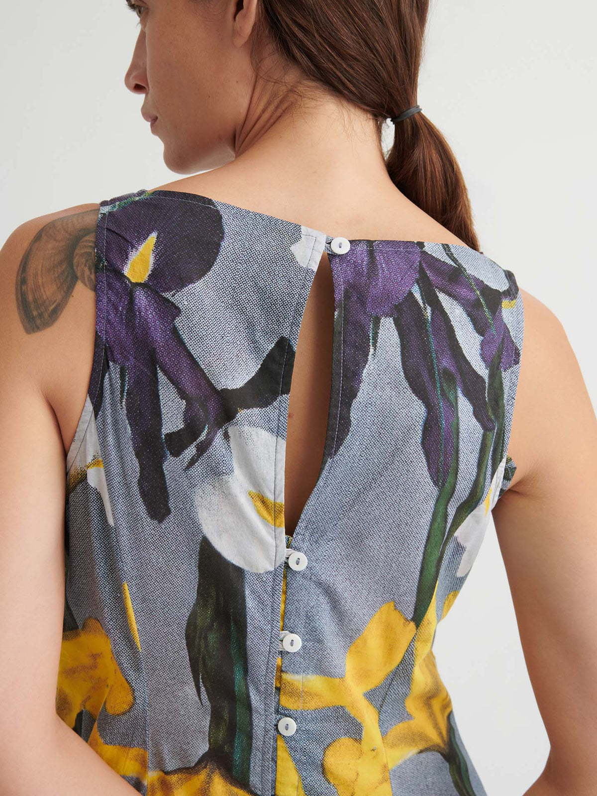 A person with a ponytail wears the Signify Dress – Iris—a sleeveless, floral dress in a standard women’s fit with back button closure and a shoulder tattoo visible.