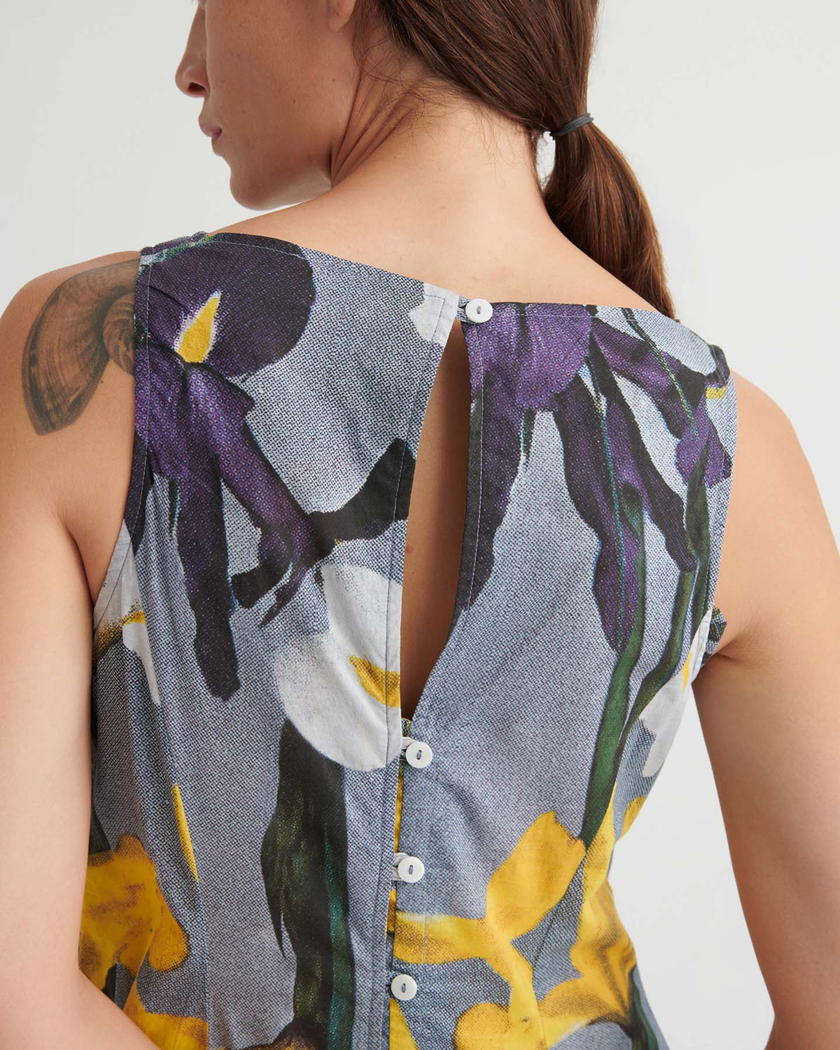 A person with a ponytail wears the Signify Dress – Iris—a sleeveless, floral dress in a standard women’s fit with back button closure and a shoulder tattoo visible.