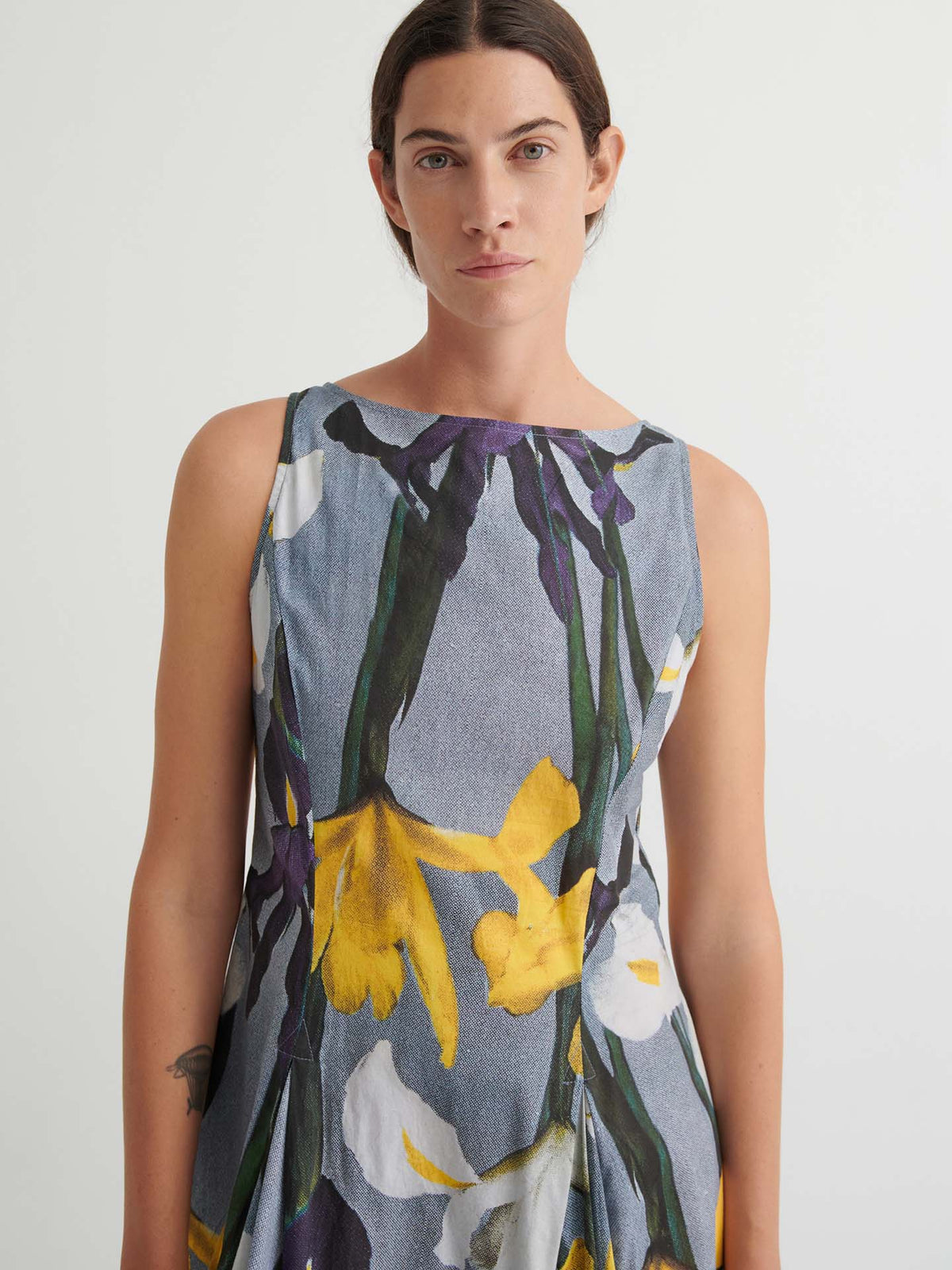 Person wearing the Signify Dress – Iris, a sleeveless garment featuring an iris-inspired floral print in yellow, white, and green hues with a standard fit, standing against a plain light background.