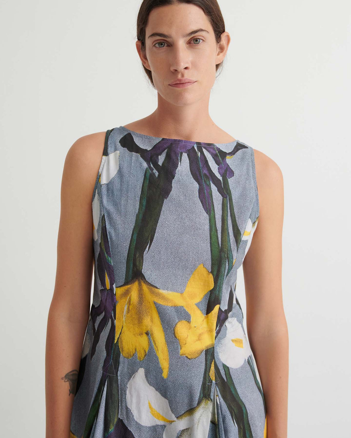 Person wearing the Signify Dress – Iris, a sleeveless garment featuring an iris-inspired floral print in yellow, white, and green hues with a standard fit, standing against a plain light background.