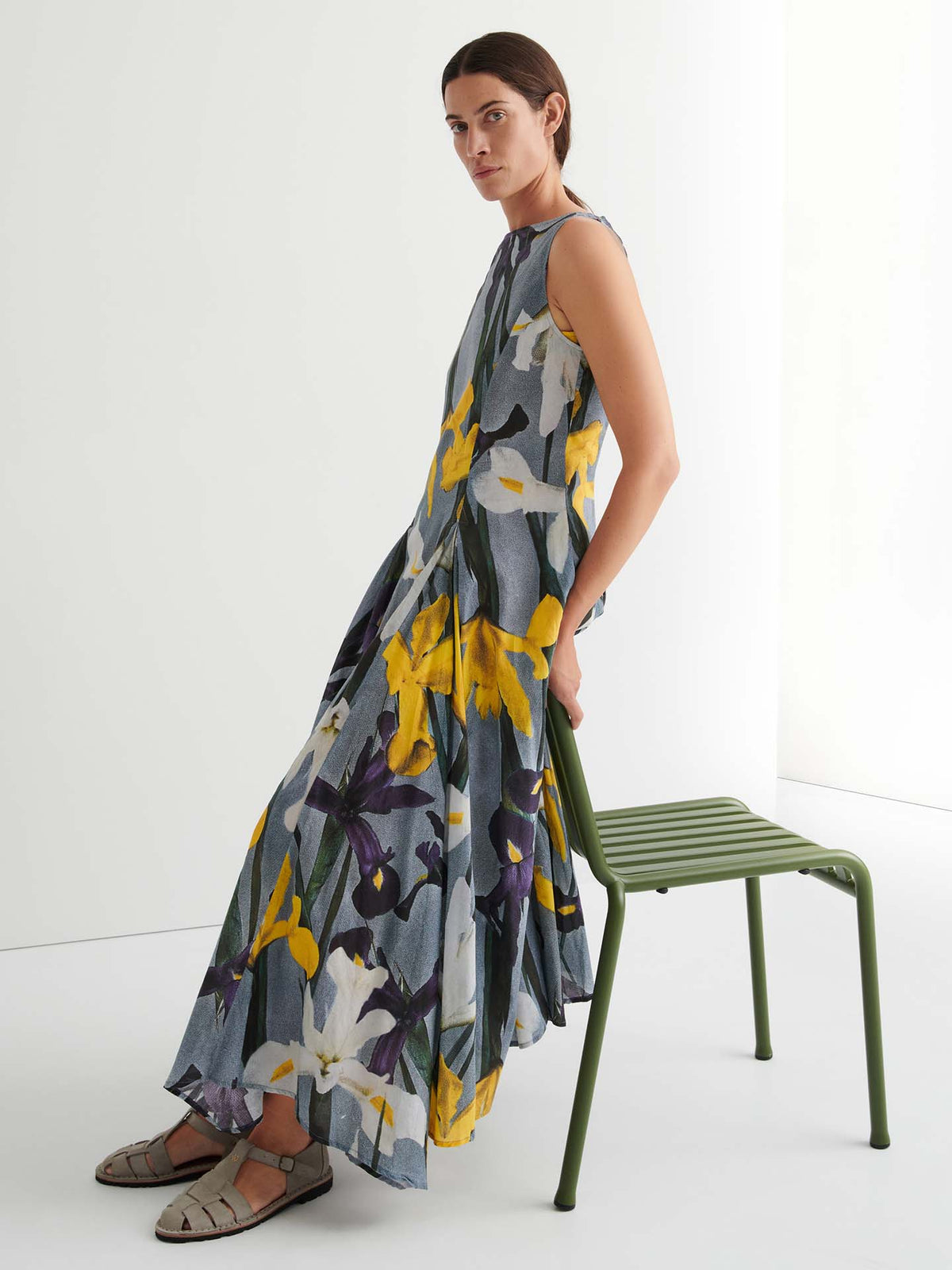A person in a sleeveless, floral-print Signify Dress – Iris with a standard fit and sandals sits sideways on a green metal chair against a plain white background.