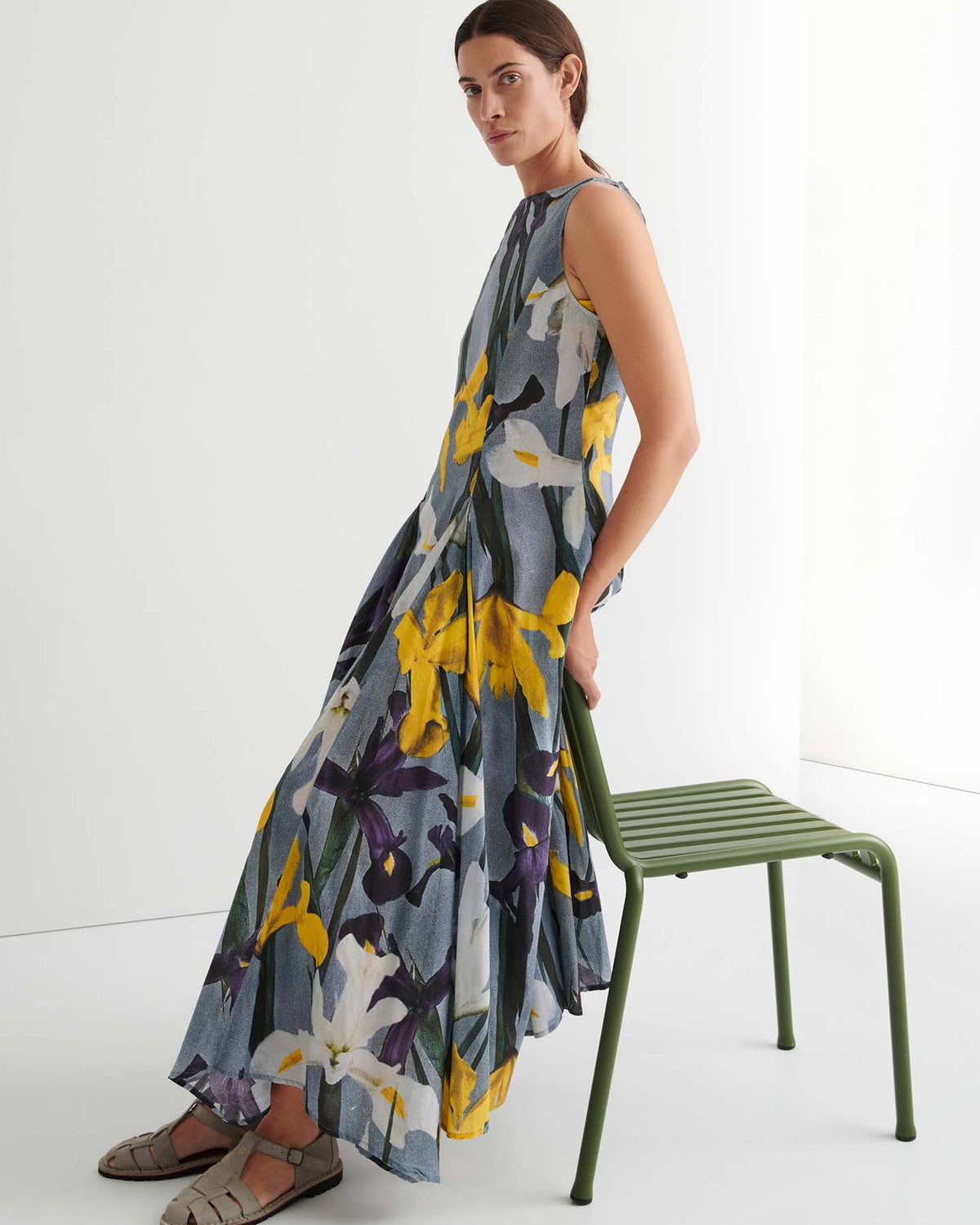 A person in a sleeveless, floral-print Signify Dress – Iris with a standard fit and sandals sits sideways on a green metal chair against a plain white background.