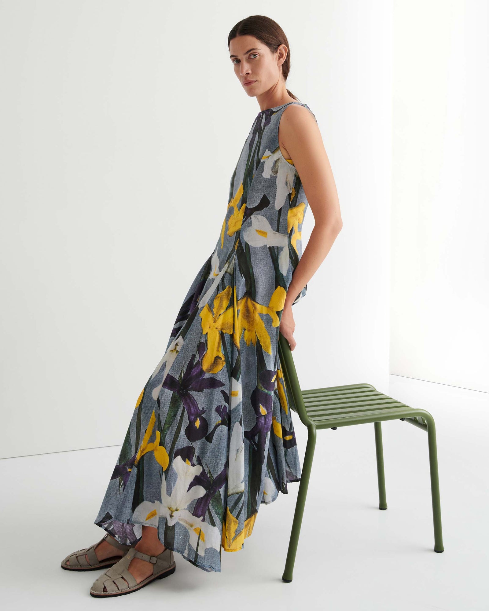 Person standing against a plain white background wearing the Signify Dress – Iris, a sleeveless, ankle-length piece featuring a bold yellow, white, and purple floral pattern. The garment’s standard fit offers ease of movement and pairs perfectly with flat sandals.