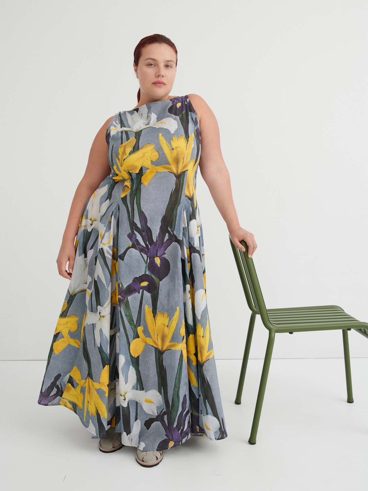 A person wearing the sleeveless, floor-length Signify Dress – Iris, featuring a yellow and white floral pattern, stands beside a green chair against a white background. The standard fit of the dress ensures comfort and elegance for any occasion.