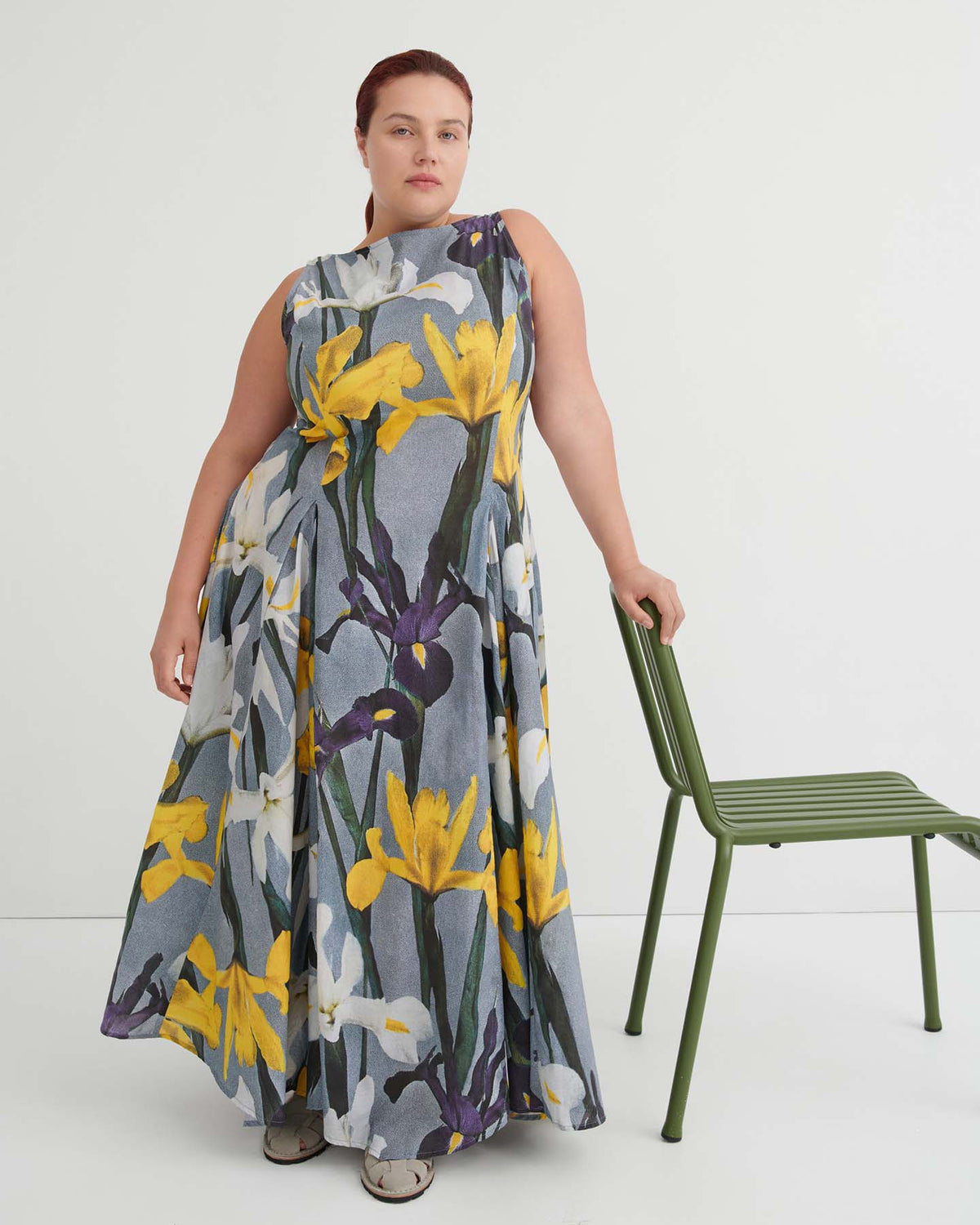 A person wearing the sleeveless, floor-length Signify Dress – Iris, featuring a yellow and white floral pattern, stands beside a green chair against a white background. The standard fit of the dress ensures comfort and elegance for any occasion.