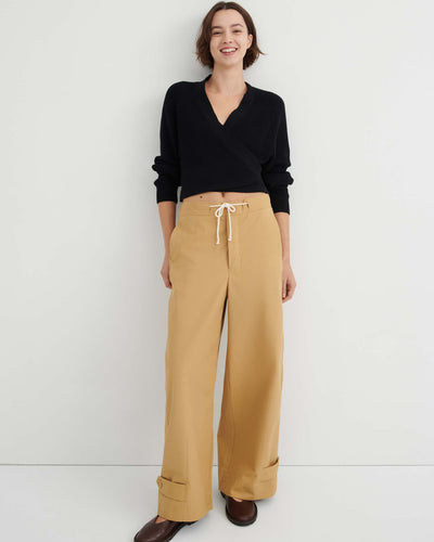 A woman stands against a white wall, wearing a black long-sleeve wrap top, the Tab Trouser in Driftwood—a relaxed fit with a natural waist and drawstring in tan—and brown shoes.