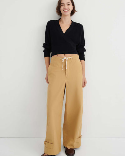 A woman stands against a white wall, wearing a black long-sleeve wrap top, the Tab Trouser in Driftwood—a relaxed fit with a natural waist and drawstring in tan—and brown shoes.