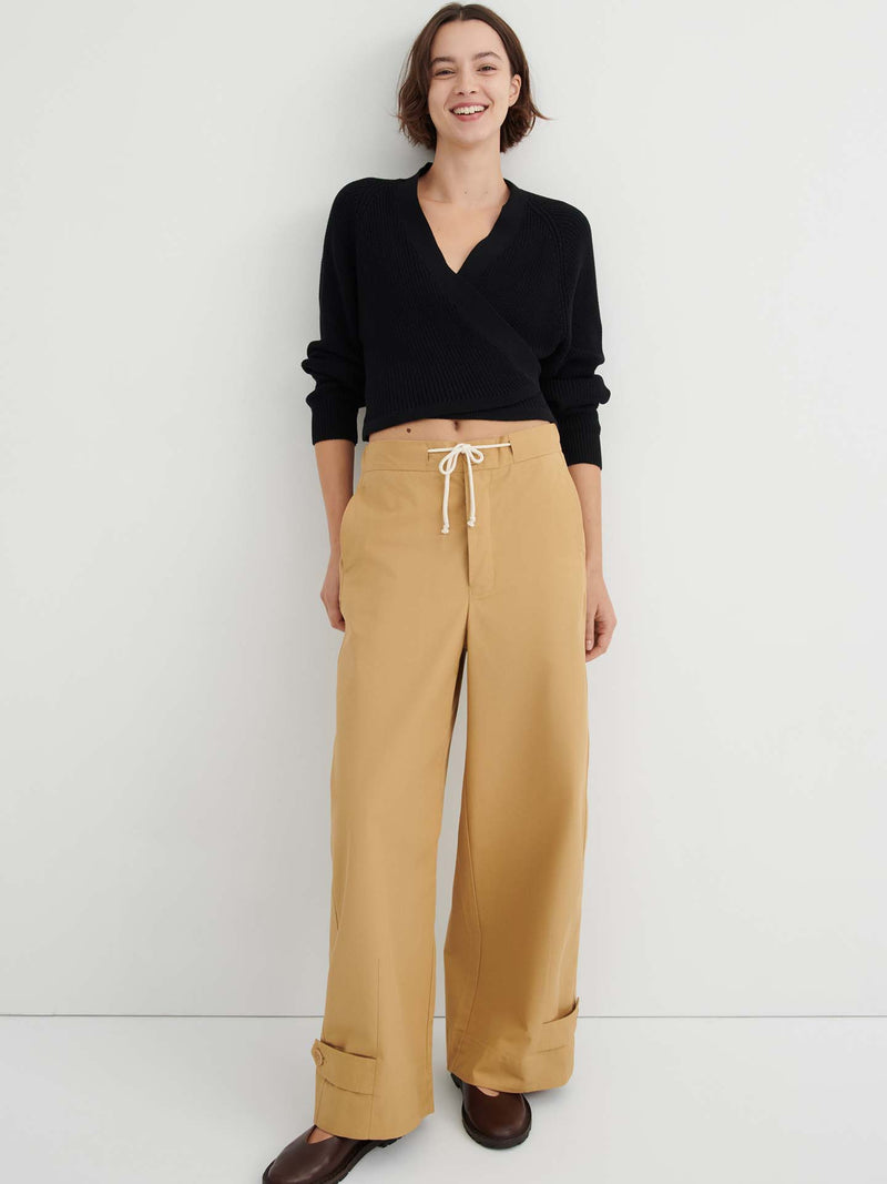 A woman stands against a white wall, wearing a black long-sleeve wrap top, the Tab Trouser in Driftwood—a relaxed fit with a natural waist and drawstring in tan—and brown shoes.