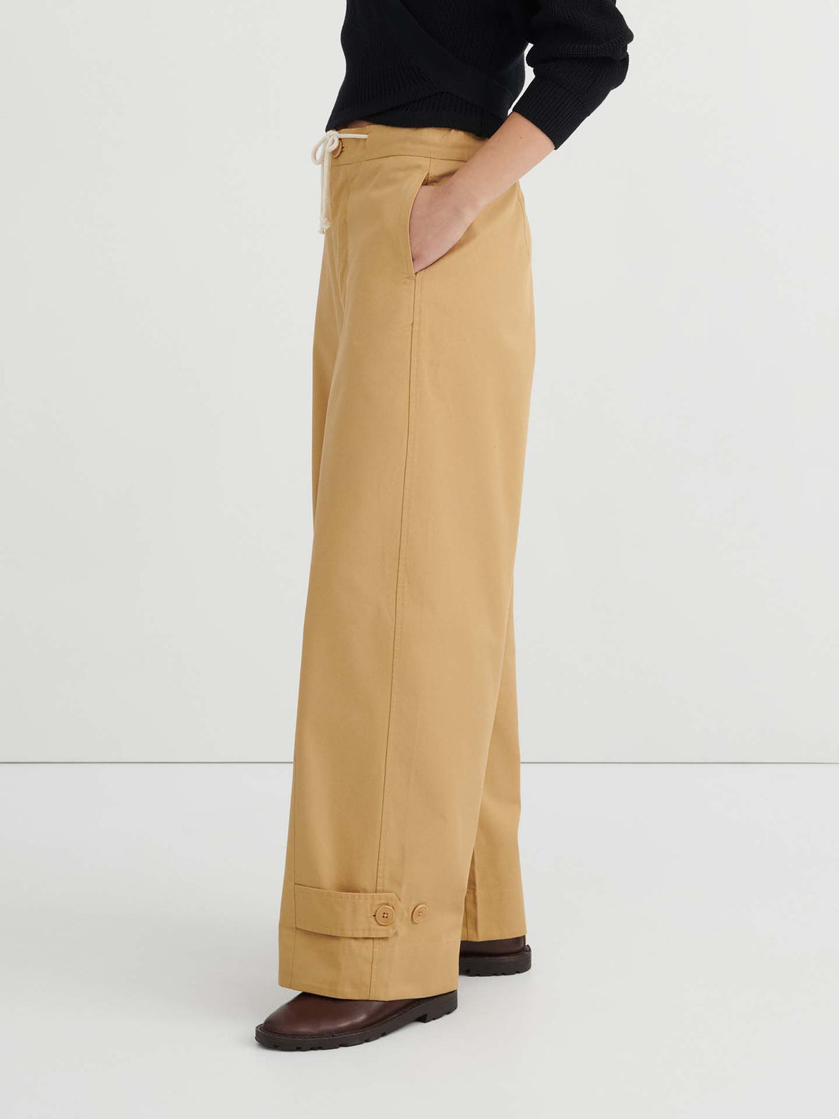 A person is wearing the Tab Trouser – Driftwood, featuring a relaxed fit with cuffed hems that sit at the natural waist, styled with a black long-sleeve top and brown shoes, standing against a plain white background.