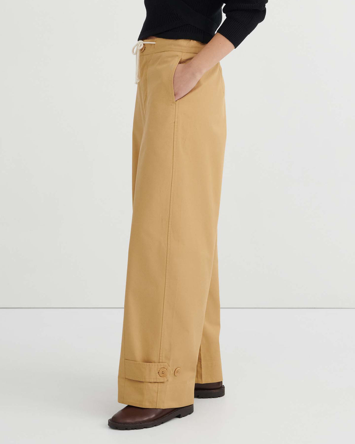 A person is wearing the Tab Trouser – Driftwood, featuring a relaxed fit with cuffed hems that sit at the natural waist, styled with a black long-sleeve top and brown shoes, standing against a plain white background.