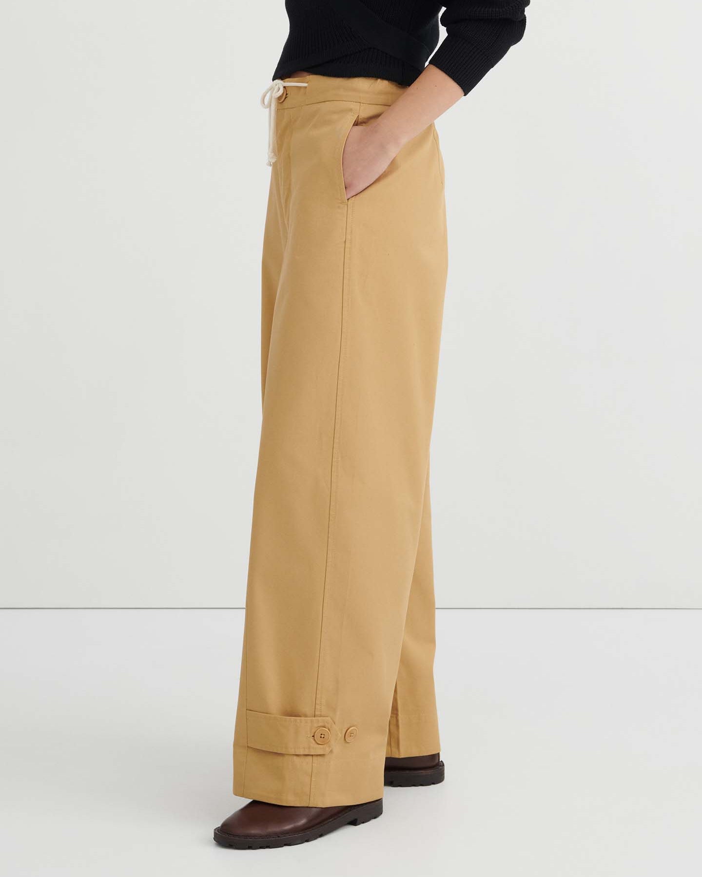 A woman stands against a white wall, wearing a black long-sleeve wrap top, the Tab Trouser in Driftwood—a relaxed fit with a natural waist and drawstring in tan—and brown shoes.