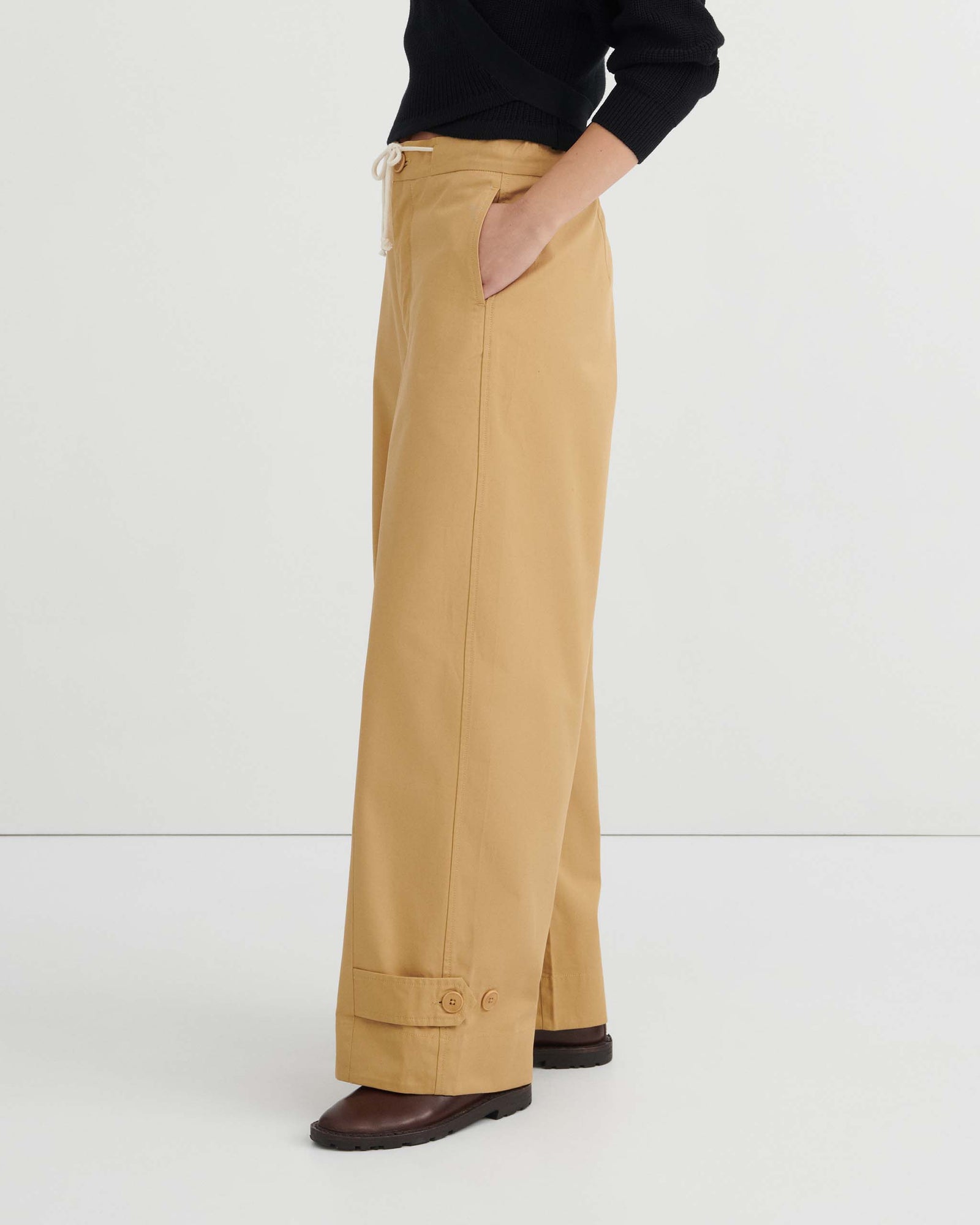 A woman stands against a white wall, wearing a black long-sleeve wrap top, the Tab Trouser in Driftwood—a relaxed fit with a natural waist and drawstring in tan—and brown shoes.