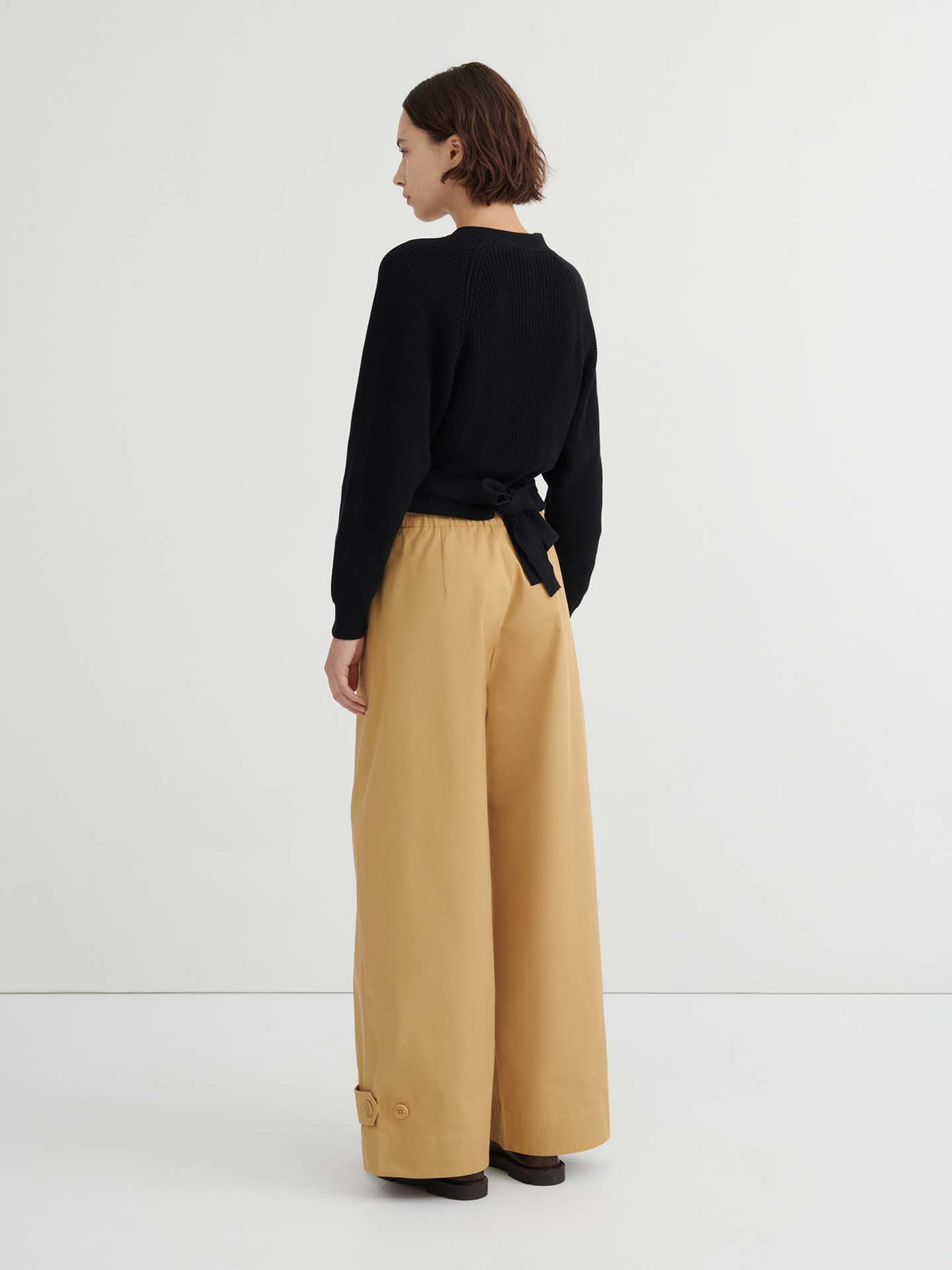 A woman stands against a plain background, seen from the back, wearing a black long-sleeve top with a tied belt and wide-leg Tab Trouser – Driftwood featuring a natural waist.
