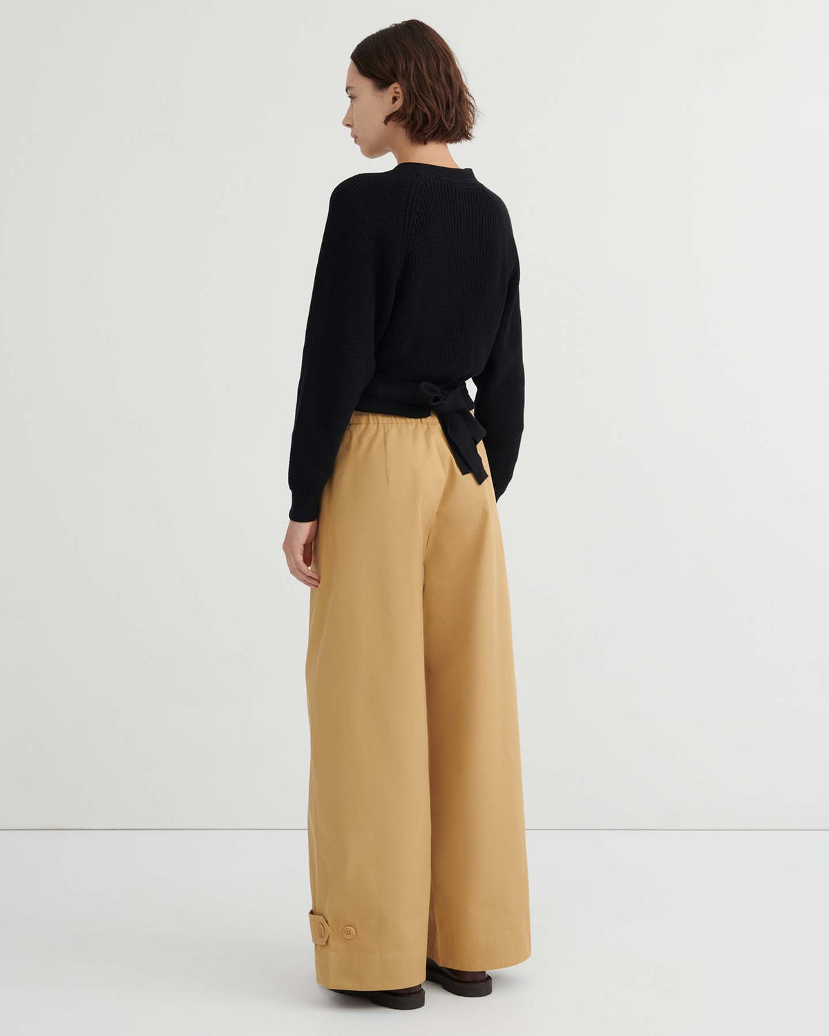 A woman stands against a plain background, seen from the back, wearing a black long-sleeve top with a tied belt and wide-leg Tab Trouser – Driftwood featuring a natural waist.