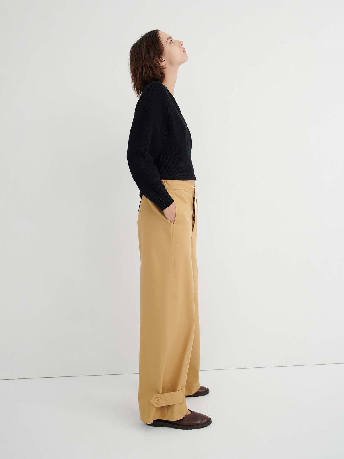 A person stands in profile against a white background, wearing a black sweater, the Tab Trouser – Driftwood with a relaxed fit, natural waist, and pockets in tan, and brown shoes, looking upwards.