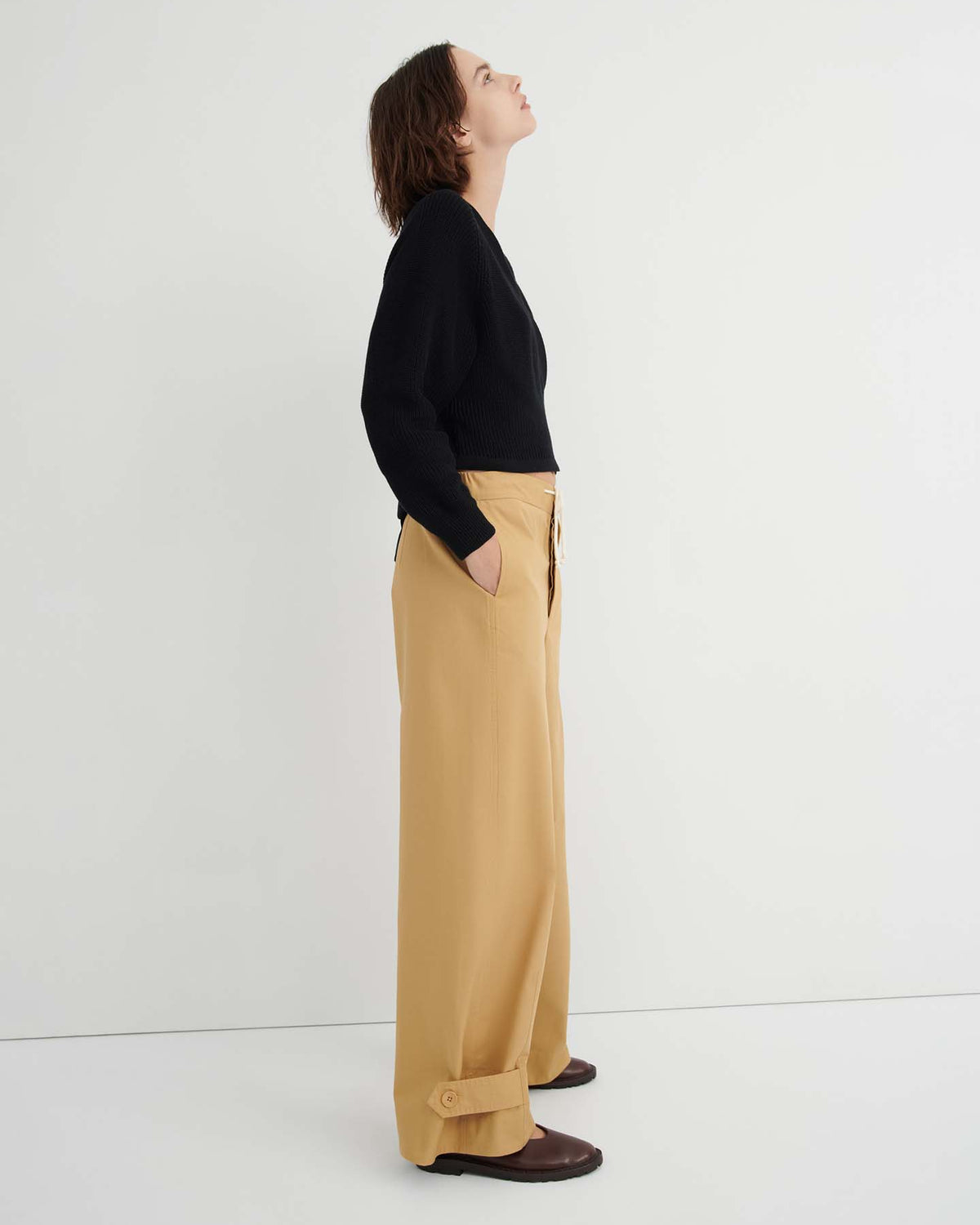 A person stands in profile against a white background, wearing a black sweater, the Tab Trouser – Driftwood with a relaxed fit, natural waist, and pockets in tan, and brown shoes, looking upwards.