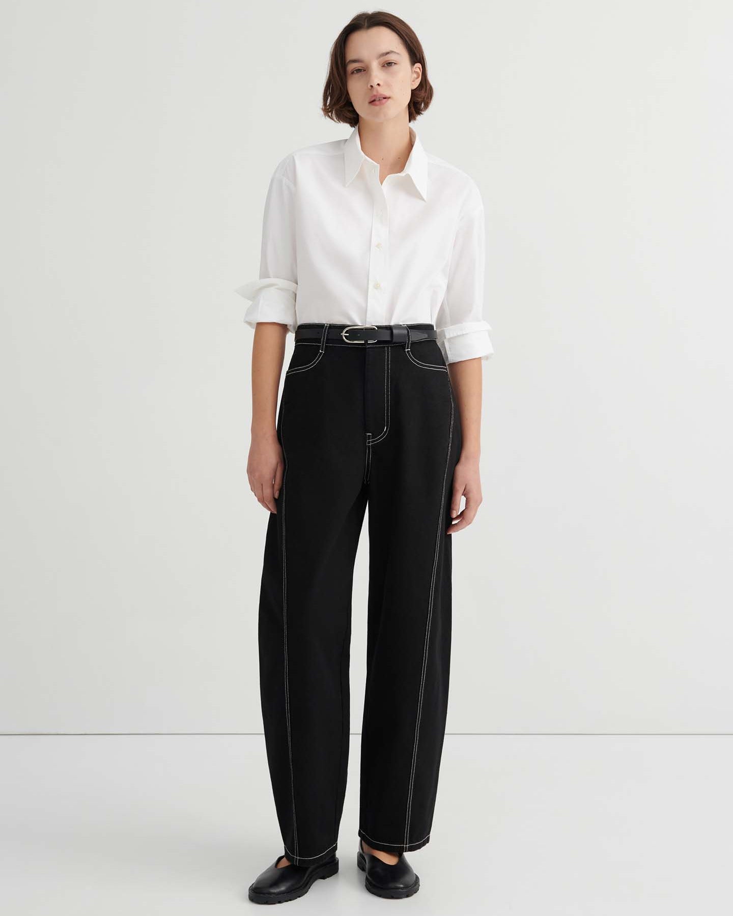 A person stands against a plain background wearing a white button-up shirt, Twist Jeans – Black with high waisted wide-leg design and white stitching, paired with a black belt and black shoes.