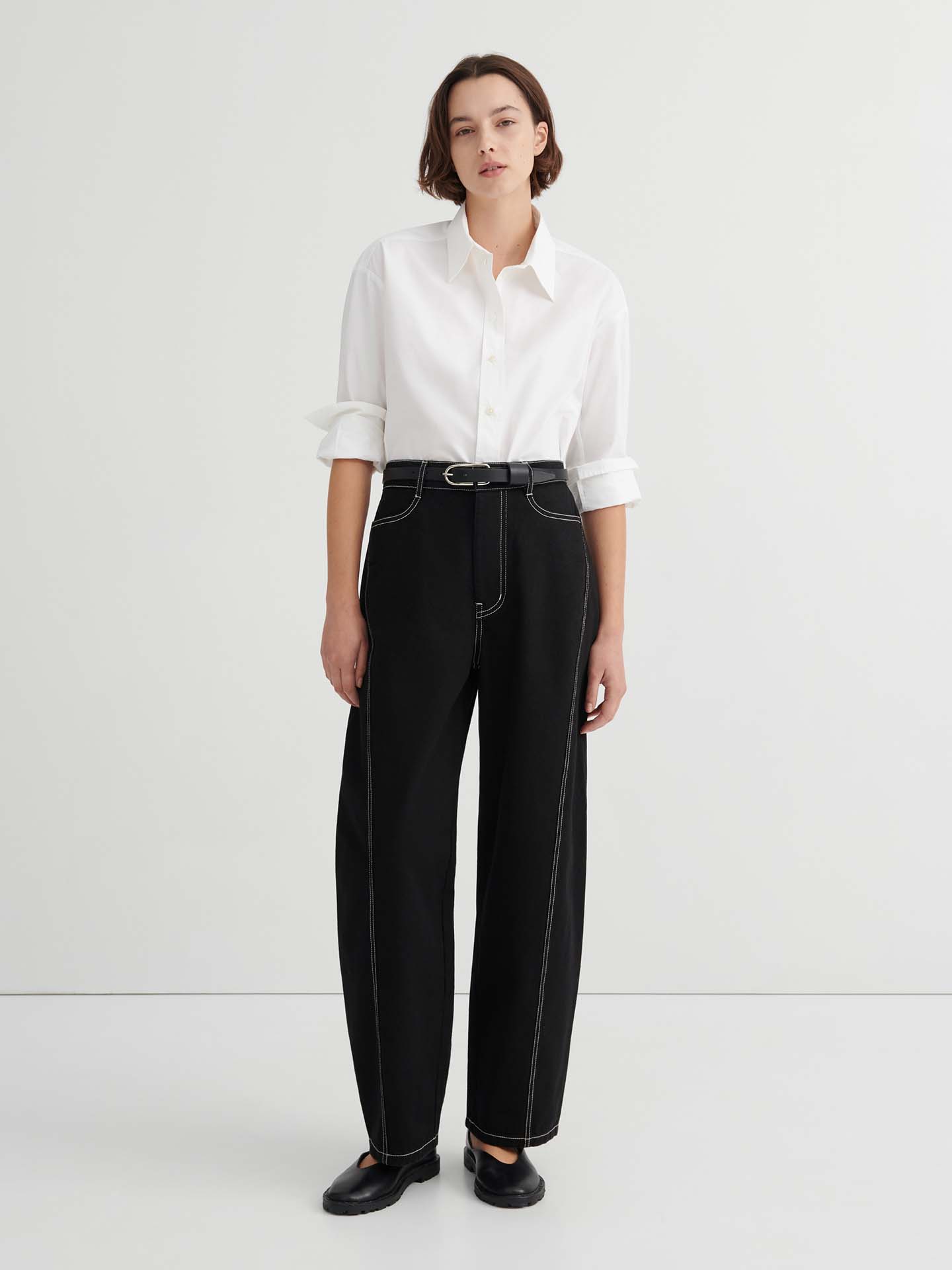 A person stands against a plain background wearing a white button-up shirt, Twist Jeans – Black with high waisted wide-leg design and white stitching, paired with a black belt and black shoes.