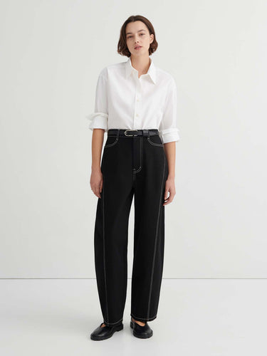 A person stands against a plain background wearing a white button-up shirt, Twist Jeans – Black with high waisted wide-leg design and white stitching, paired with a black belt and black shoes.