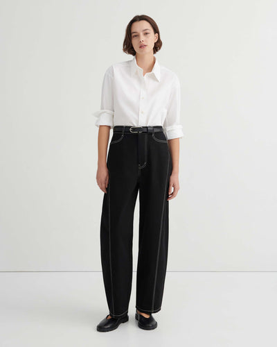 A person stands against a plain background wearing a white button-up shirt, Twist Jeans – Black with high waisted wide-leg design and white stitching, paired with a black belt and black shoes.