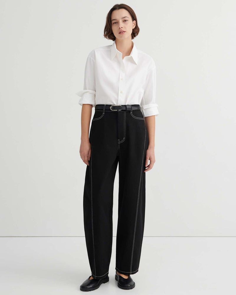 A person stands against a plain background wearing a white button-up shirt, Twist Jeans – Black with high waisted wide-leg design and white stitching, paired with a black belt and black shoes.