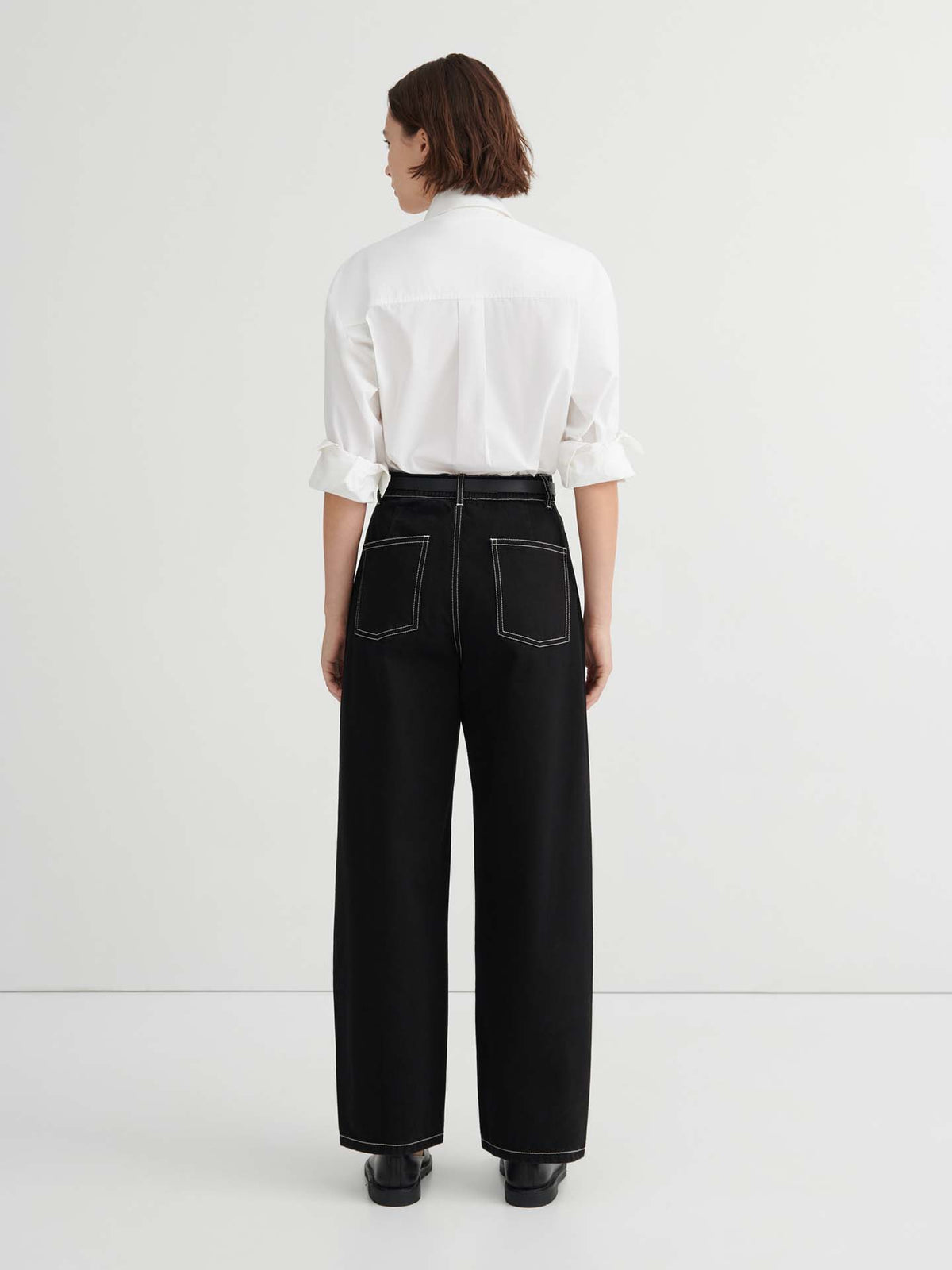 A person stands with their back to the camera in a plain white room, wearing a white long-sleeve shirt and high-waisted, relaxed fit Twist Jeans – Black, featuring white stitching.