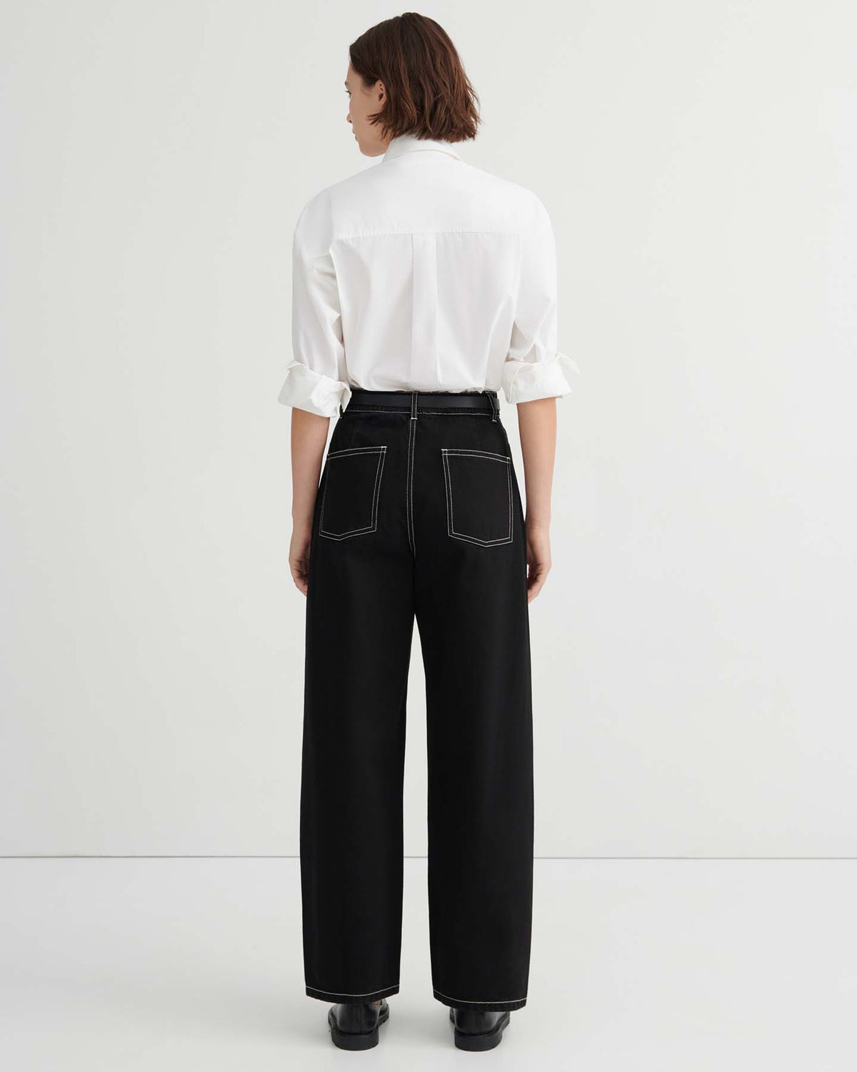A person stands with their back to the camera in a plain white room, wearing a white long-sleeve shirt and high-waisted, relaxed fit Twist Jeans – Black, featuring white stitching.