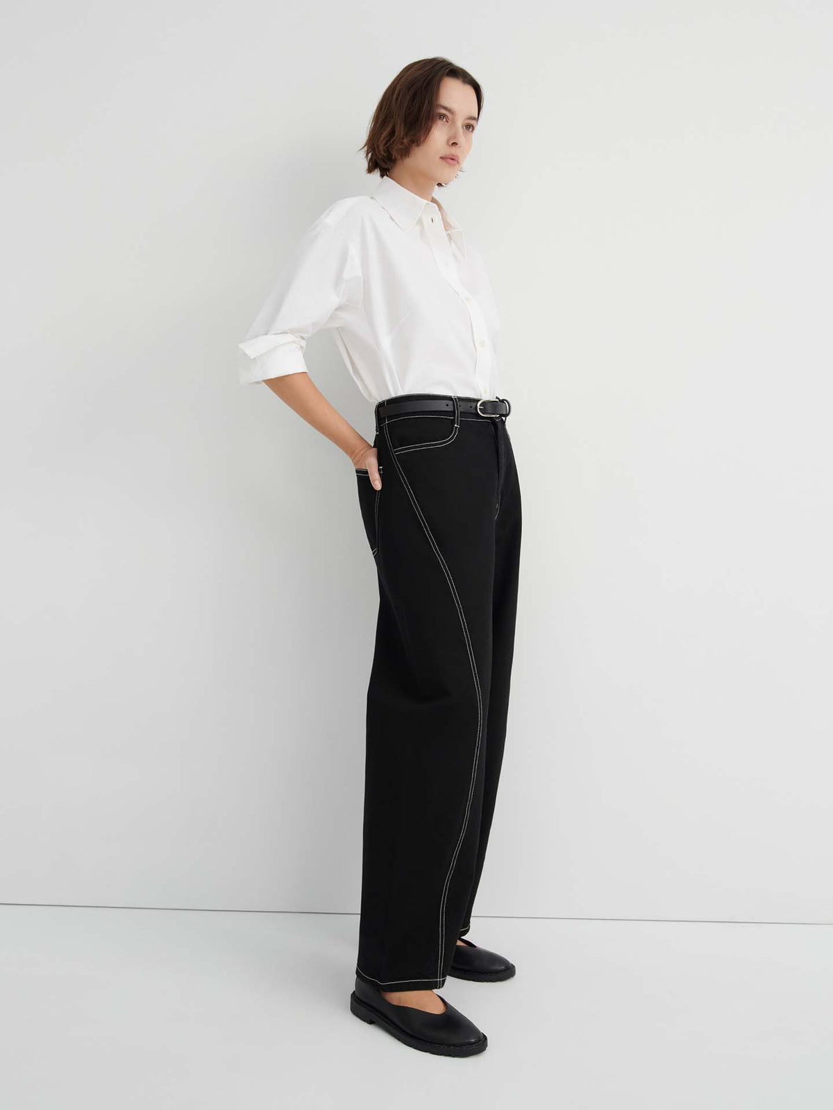 A person stands against a plain white background wearing a white button-up shirt, Twist Jeans – Black in a relaxed, high-waisted wide-leg fit with white stitching, and black shoes.