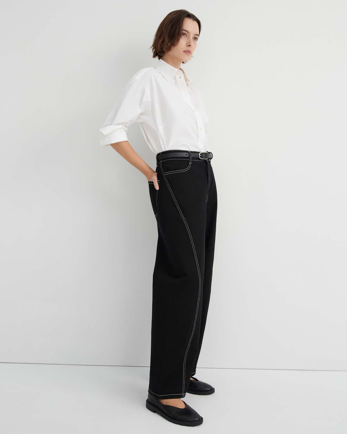 A person stands against a plain white background wearing a white button-up shirt, Twist Jeans – Black in a relaxed, high-waisted wide-leg fit with white stitching, and black shoes.