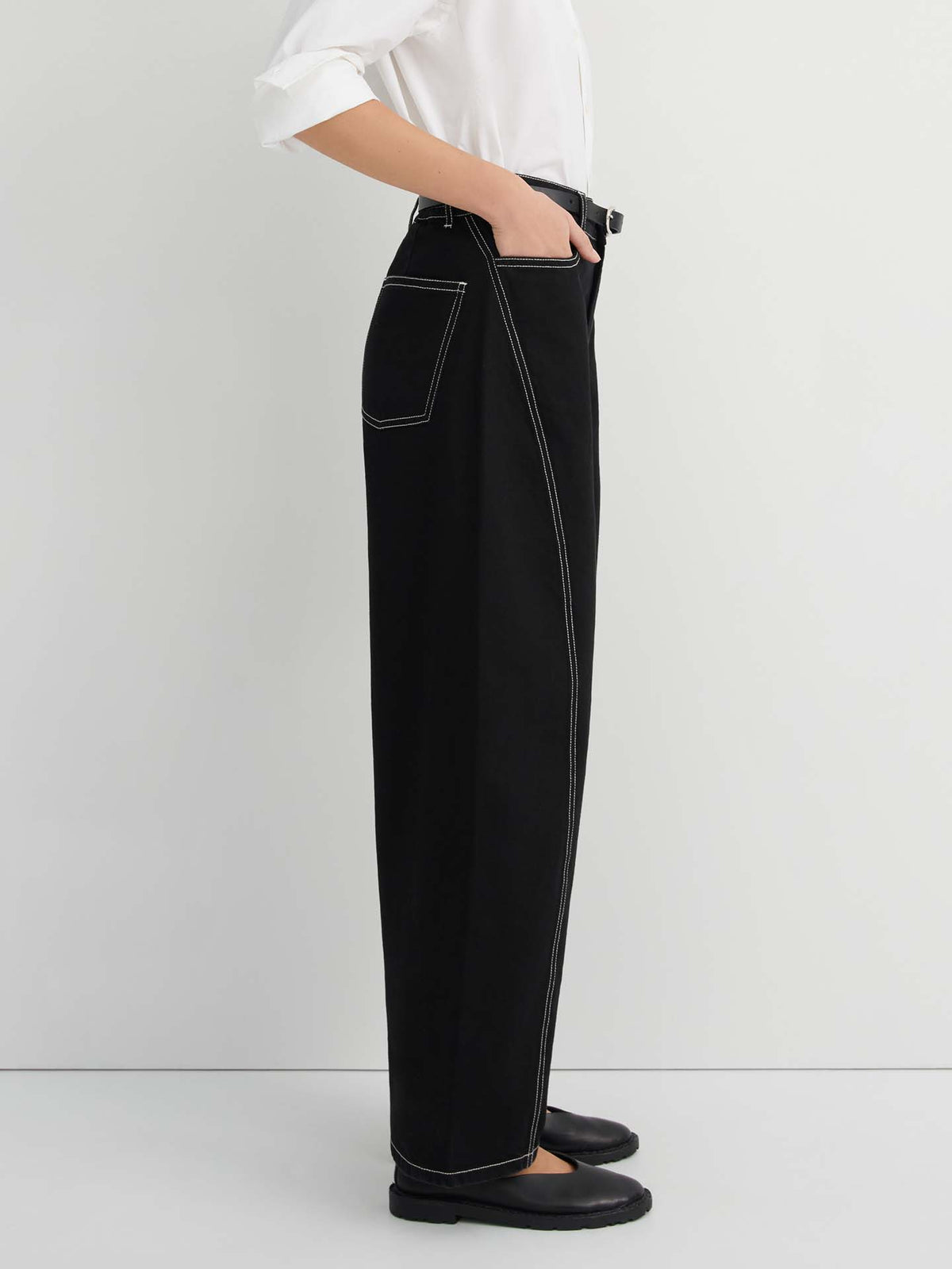 A person is standing sideways against a plain light background, wearing Twist Jeans – Black styled in a high-waisted, wide-leg silhouette with white contrast stitching, paired with a white shirt and black shoes, highlighting the relaxed fit of the jeans.