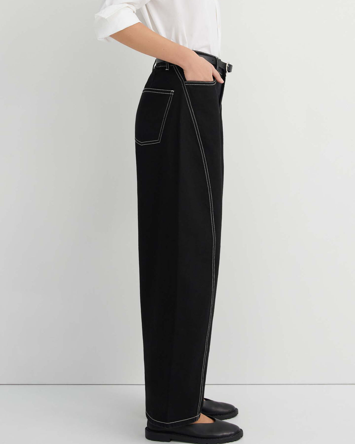 A person is standing sideways against a plain light background, wearing Twist Jeans – Black styled in a high-waisted, wide-leg silhouette with white contrast stitching, paired with a white shirt and black shoes, highlighting the relaxed fit of the jeans.