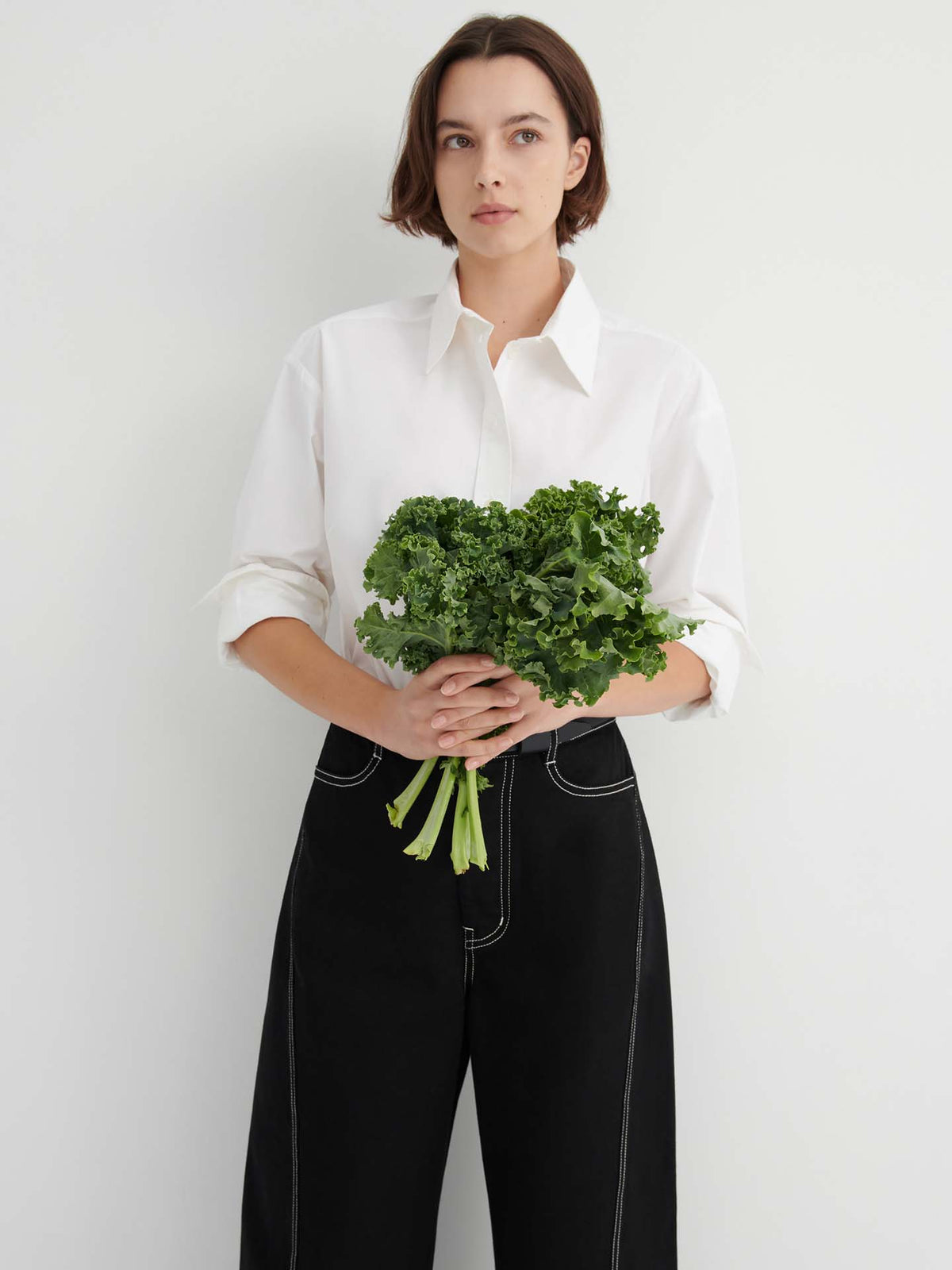 A person stands against a white wall, wearing a white shirt and the Twist Jeans – Black, holding a bunch of leafy green kale with both hands.