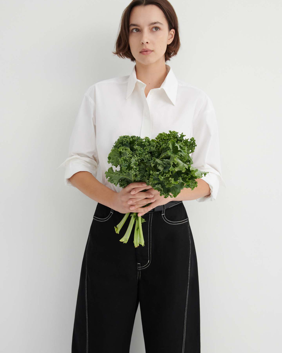 A person stands against a white wall, wearing a white shirt and the Twist Jeans – Black, holding a bunch of leafy green kale with both hands.